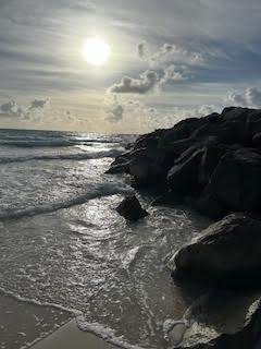 Waves wash against large rocks on a sandy beach under a cloudy sky with the sun shining above the horizon.