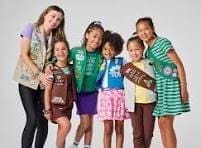 A group of six girls wearing different Girl Scouts uniforms stand together and smile in front of a plain background.