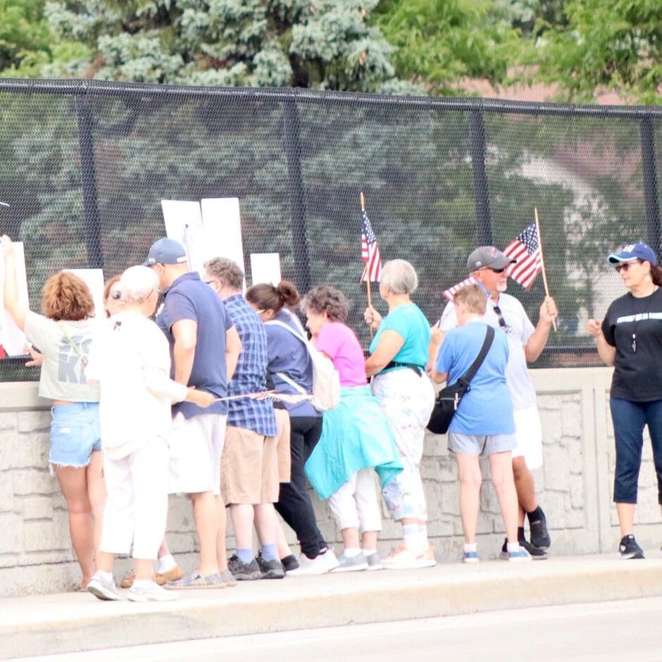 A group of people stand along a sidewalk next to a fence, some holding small American flags and signs.
