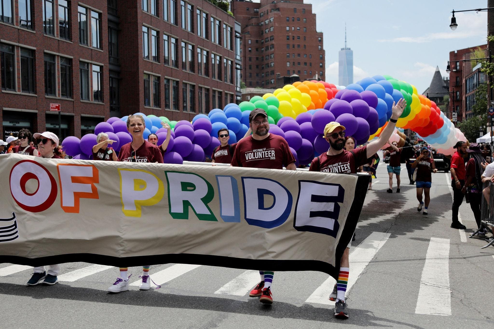 People march in a Pride parade, holding a banner and walking under a large rainbow balloon arch on a city street.