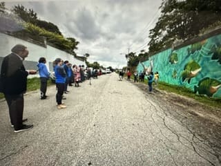 People stand in two lines along a cracked street, some looking at a mural of fish painted on a wall to the right, under a cloudy sky.