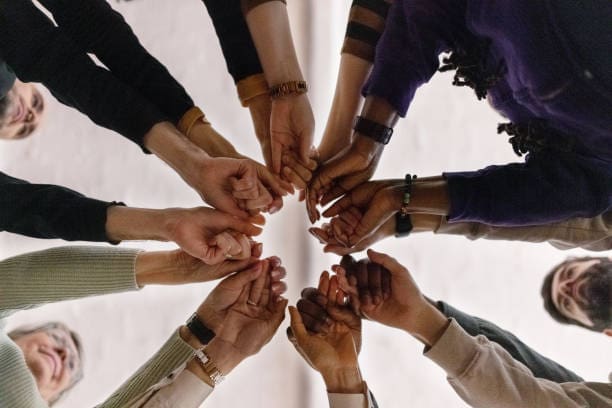 A group of people stand in a circle with their hands extended toward the center, fingers interlocked.