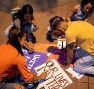 Four people gather on the ground, creating and decorating protest signs with messages such as "OUR LIVES MATTER.