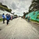 People stand in two lines along a cracked street, some looking at a mural of fish painted on a wall to the right, under a cloudy sky.