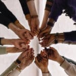 A group of people stand in a circle with their hands extended toward the center, fingers interlocked.